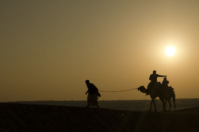 Silhouette man pulling camels on desert against sky during sunset