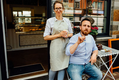 Man and woman smiling at camera while drinking tea at entrance of cafe