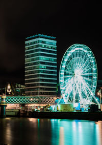 Illuminated ferris wheel by river against sky in city at night