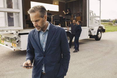 Businessman using smart phone with colleagues and portable office truck on road in background