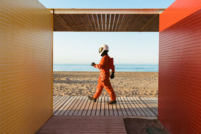 Side view of astronaut in spacesuit and helmet walking by futuristic building and admiring sea against blue sky on alien planet