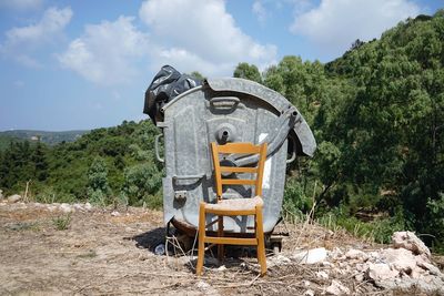 Old chair in front of bin on field by trees against sky