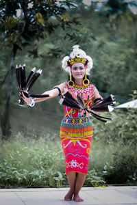 Portrait of young woman with arms raised standing against trees