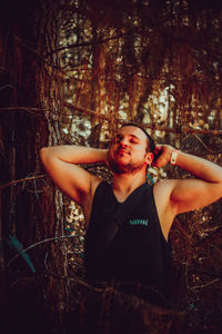 Man with closed eyes standing by tree trunk in forest