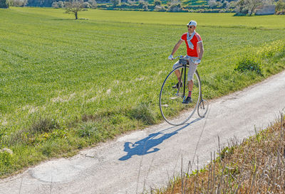 From above adult focused male in sunglasses and stylish casual clothes riding retro high wheel bike along footpath against green grassy field in summer day