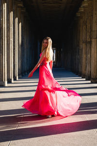 Full length portrait of young woman in pink evening gown standing at colonnade