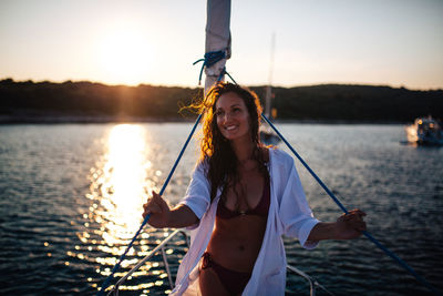 Thoughtful smiling woman looking away while standing on boat in lake during sunset