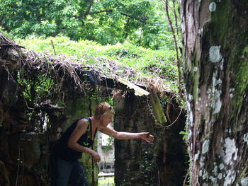 Side view of woman looking at plant while standing in forest