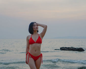 Woman with hand in hair wearing bikini standing on beach against sky during sunset
