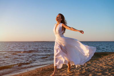 Young woman in summer in a white dress on the shore at sunset