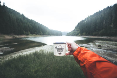 Cropped hand holding mug at lakeshore against mountains