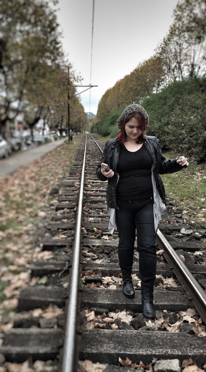 railroad track, casual clothing, lifestyles, rail transportation, full length, person, looking at camera, standing, portrait, young adult, transportation, front view, leisure activity, warm clothing, tree, day, jacket, the way forward