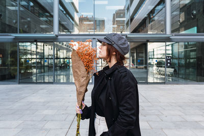 Side view of young woman in stylish outerwear with cap and glasses closing eyes and enjoying smell of orange flowers against modern building with glass walls on city street