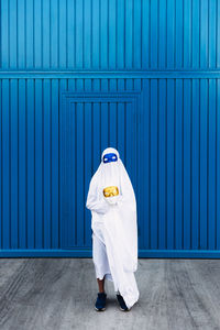 Unrecognizable kid wearing masquerade mask and ghost costume standing in city during halloween and looking at camera