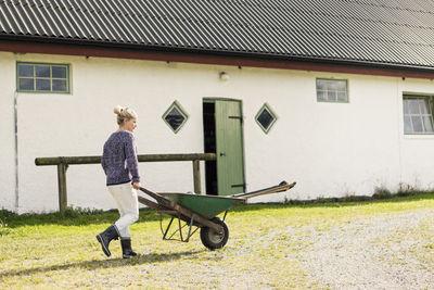 Female farm worker walking with wheelbarrow on road by barn