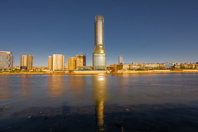 Buildings by river against clear blue sky