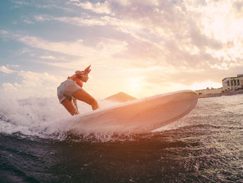 Low angle view of young woman surfing on sea against sky during sunset