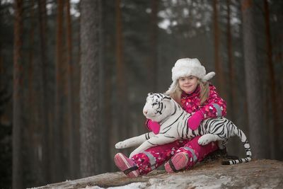 Portrait of girl in warm clothing holding toy tiger while sitting by trees in forest