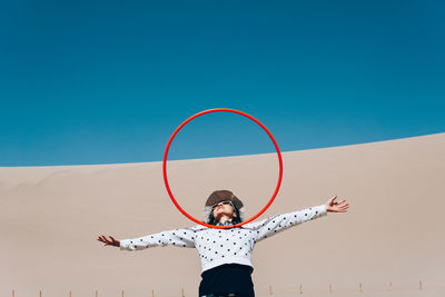 Woman balancing hoop while standing against wall and clear blue sky