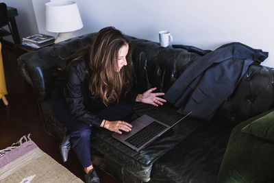 High angle of young female entrepreneur in formal blazer sitting on sofa and communicating with partner via video call on laptop while working remotely from home