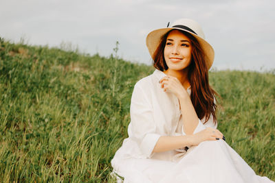 Woman looking away while sitting on grass against sky