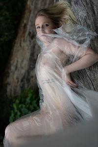 Portrait of naked woman wrapped in plastic standing by tree trunk