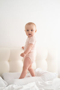 Cute baby girl in bodysuit stand holding bed frame, learning to walk on white bedding on bed