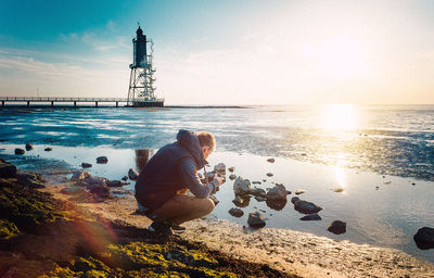 Man photographing through camera by sea against sky during sunset