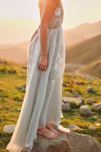 Midsection of woman standing on rock against sky during sunset