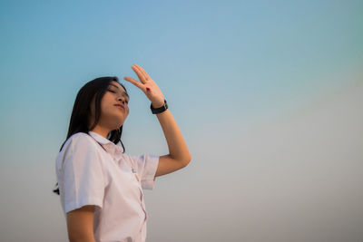 Low angle view of young woman shielding eyes standing against sky during sunset