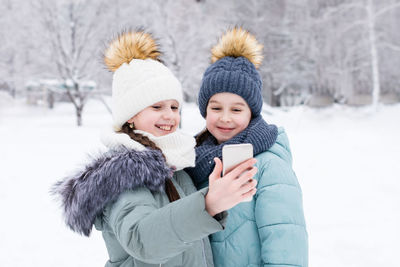 Two smiling girls in warm clothes make a video call on a smartphone in a snowy winter park. 