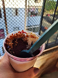 Açaí cup in one hand with paçoca and powdered milk in front of a window in the middle of a sunny day