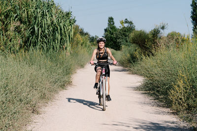 Full body of positive young female cyclist in sportswear and helmet riding bicycle on dirt path among green plants in summer day in countryside