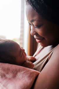 Side view of african american female with long hair standing at window and kissing newborn baby wrapped in soft towel