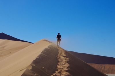 Rear view of man walking in desert against clear blue sky