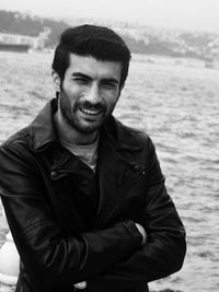 Portrait of handsome man wearing leather jacket with arms crossed against sea