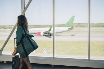 Businesswoman walking with luggage by window in corridor at airport