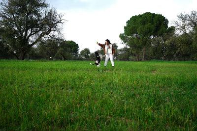 Young lady in casual outfit standing on green grass and trowing ball to black and white obedient spaniel in summer park