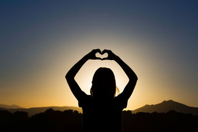 Silhouette woman standing by heart shape against sky during sunset