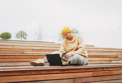 Stylish man hipster in yellow hat freelancer working on laptop in street city park