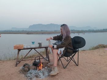 Woman sitting on chair by lake against sky