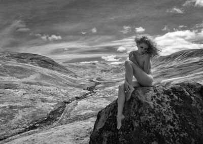 Naked young woman sitting on rock against sky