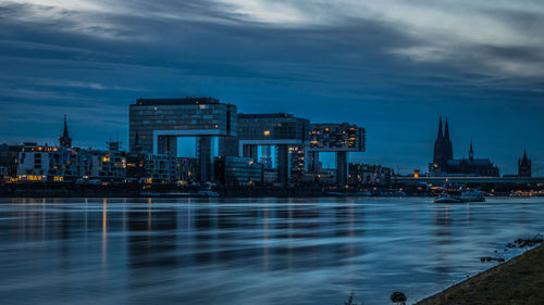 Illuminated buildings by river against cloudy sky