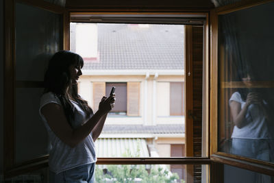 Young woman using mobile phone while standing by window at home