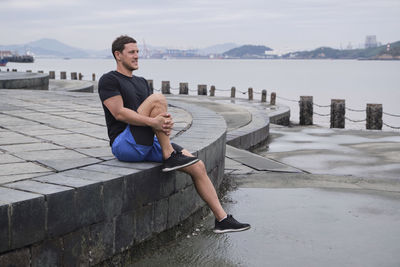 Side view of content male runner sitting on stone border and stretching legs during training on embankment in city