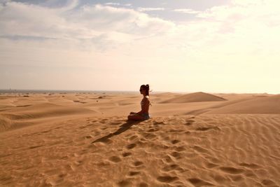 Side view of young woman sitting on sand against cloudy sky