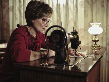 Senior lady in glasses using retro sewing machine to create linen napkin in cozy room at home