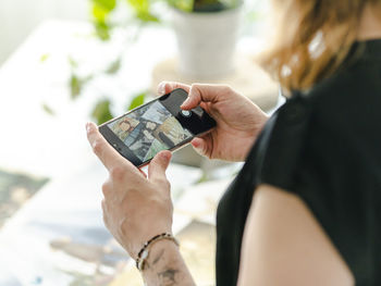 Cheerful female designer taking picture of paintings on smartphone while standing near table and working in creative workspace