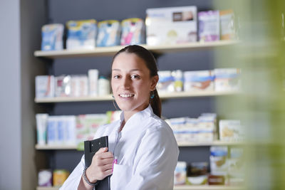 Portrait of female pharmacist with clipboard standing against shelf in store