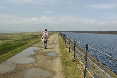 Rear view of woman walking on the dam of a reservoir in moorland against sky 
